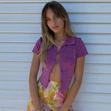 Woman wearing a purple cropped collared shirt and a floral print skirt, posing against a white slatted wall.