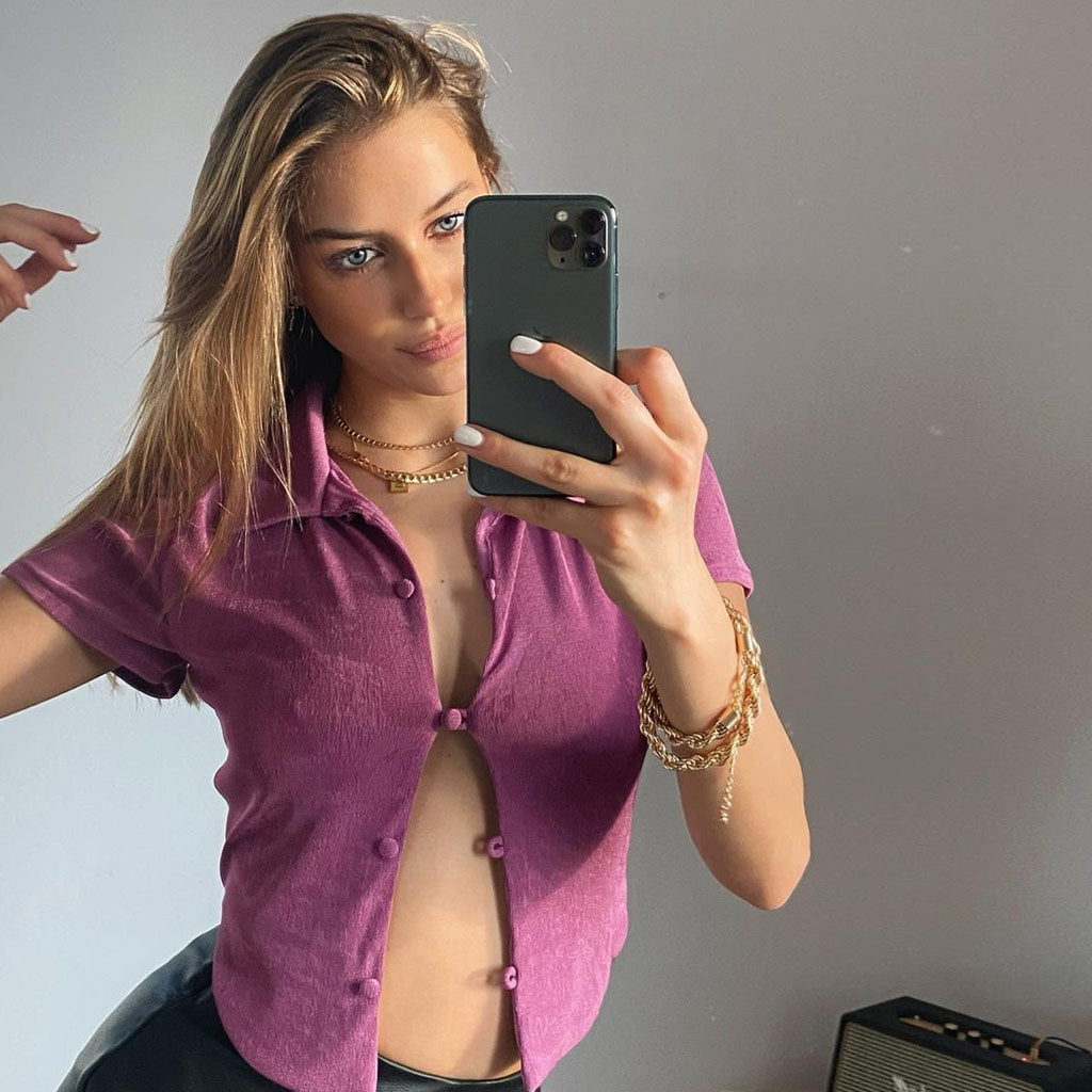 Woman taking a mirror selfie, wearing a purple cropped collared shirt open at the chest, gold necklaces, and black pants.