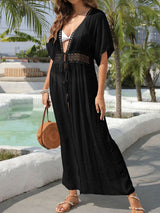 Woman wearing a black sheer maxi dress with a crochet detail at the bust and empire waist, and a high slit, standing by a pool.