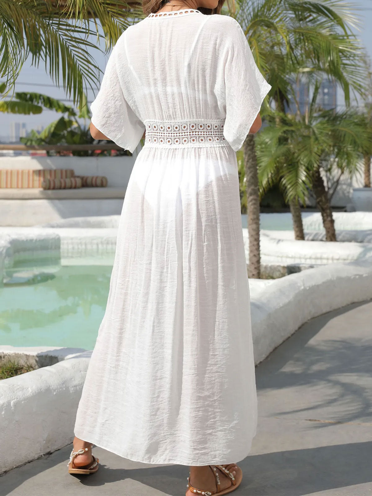 Back view of a woman wearing a white sheer maxi dress with a crochet detail at the empire waist, standing by a pool.