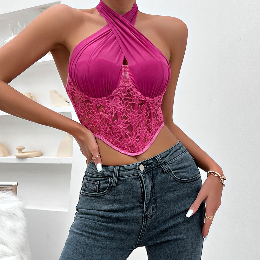 A woman wearing a pink halter top with lace detailing and blue jeans, standing in front of a white wall.