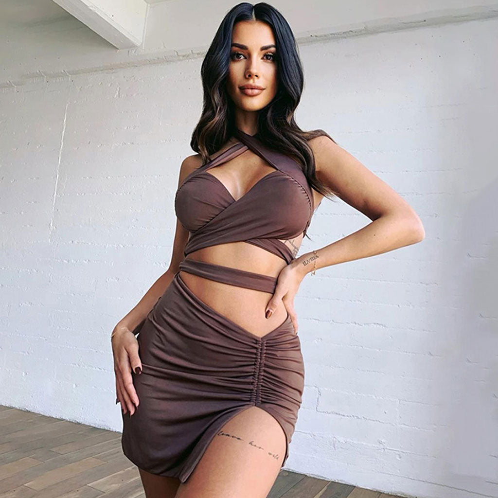 Woman wearing a brown halter neck two-piece dress with a ruched skirt and thigh-high slit, standing in front of a white brick wall.