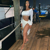 Woman in a white long-sleeved crop top and a white high-slit skirt, standing on a wooden pier at night.