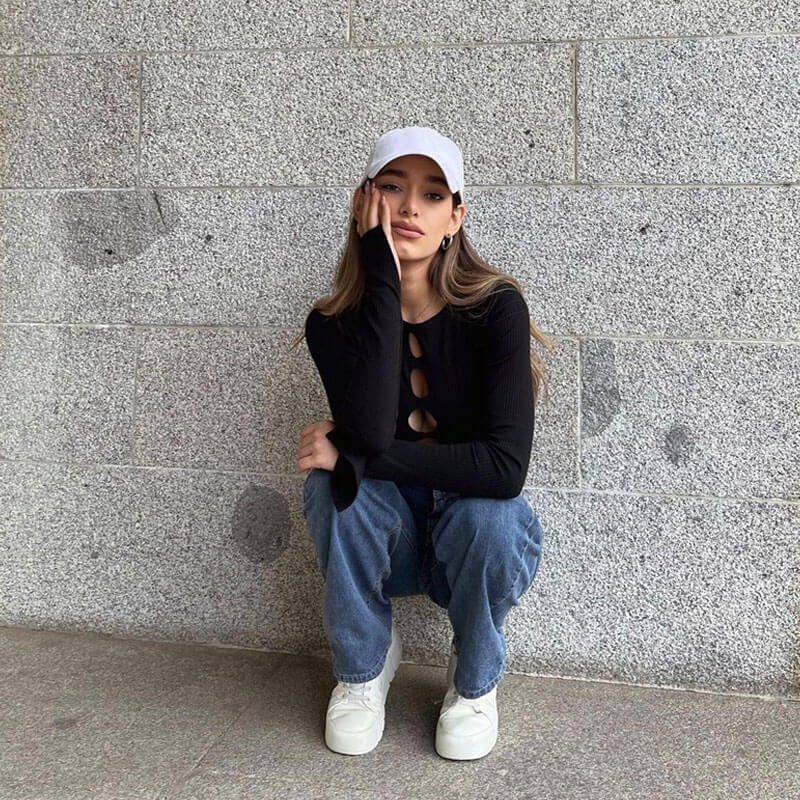 Woman wearing a black cut-out long-sleeve top and blue jeans, crouching against a stone wall, resting her hand on her face.