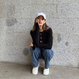 Woman wearing a black cut-out long-sleeve top and blue jeans, crouching against a stone wall, resting her hand on her face.