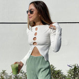 Woman in a white ribbed long-sleeve top with cut-out details and green pleated pants, holding a green drink.