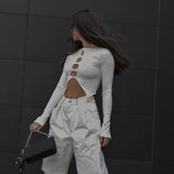Woman in a white ribbed long-sleeve top with cut-out details and white pants, holding a black purse.