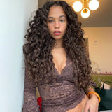 Woman with long curly brown hair wearing a brown lace top with a deep v-neckline and bell sleeves, leaning against a white wall.