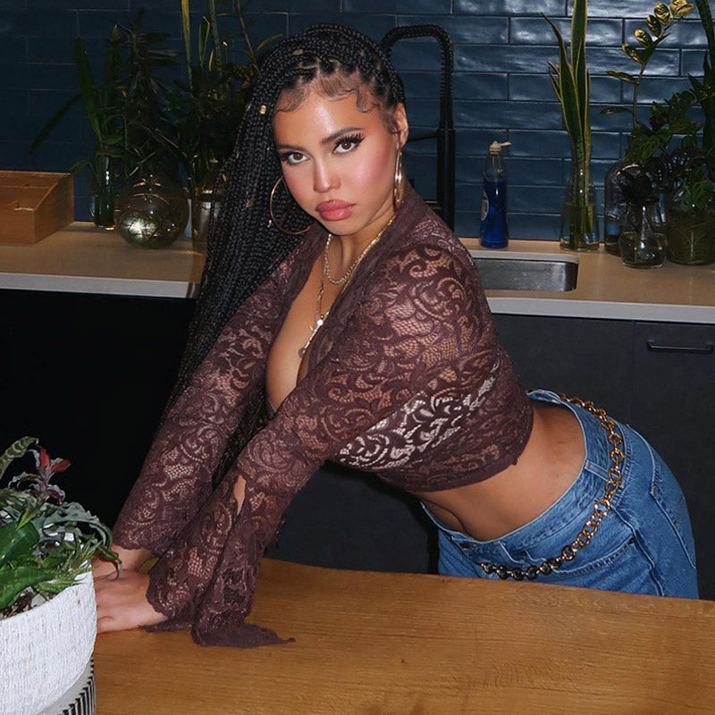 Woman with braided hair wearing a brown lace crop top with bell sleeves and blue jeans, leaning over a wooden table.