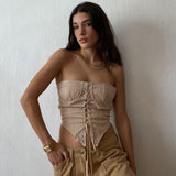 A young woman with long dark hair wearing a beige strapless corset top and tan pants, posing against a white wall.