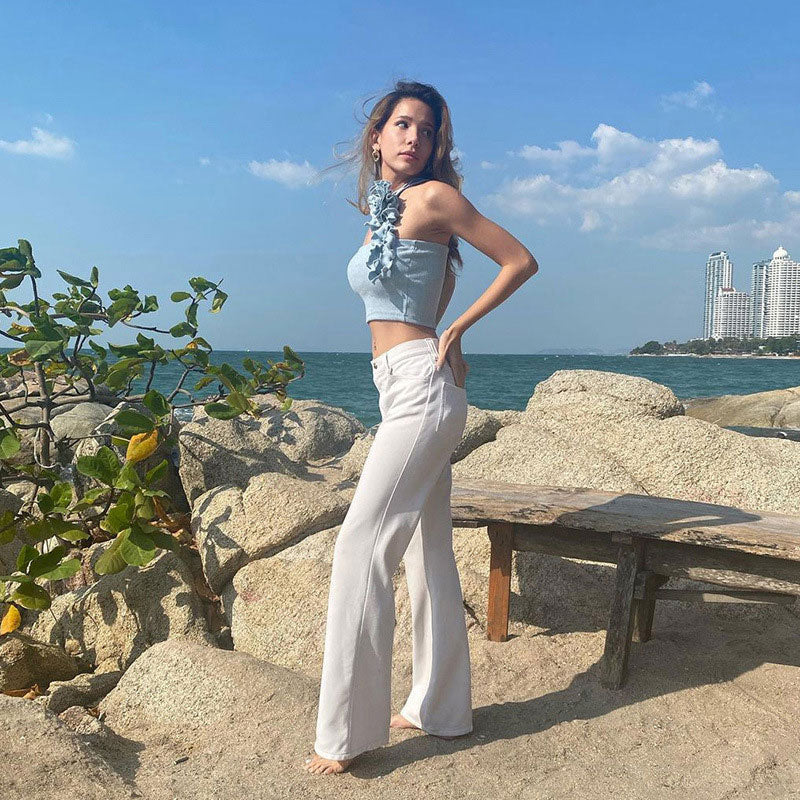 A full-body shot of a woman in a light blue crop top and white flared pants on a beach.