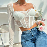 Woman wearing a white sheer long-sleeve top with a lace corset bodice and blue jeans, holding a pink rose.