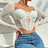 Close-up of a woman in a white sheer long-sleeve top with a lace corset bodice and blue jeans, hands on hips.