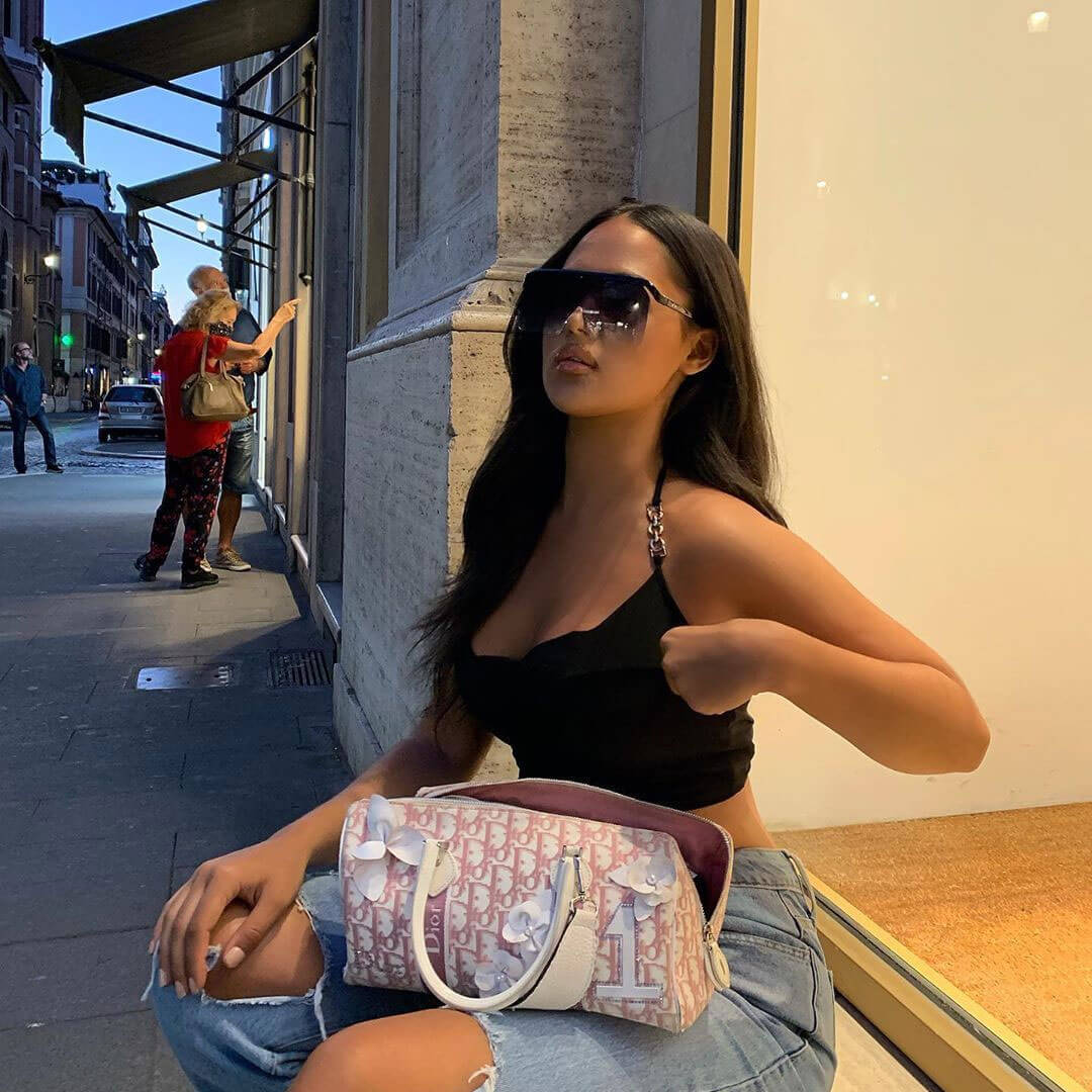 A young woman sits on the ground, wearing a black crop top, ripped jeans, and large sunglasses, with a designer handbag beside her.