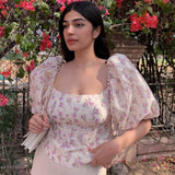 Close-up of a woman in a floral print top with puff sleeves, standing in front of pink flowers.