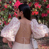 Woman in a floral print top with puff sleeves and an open back, posing in front of pink flowers.