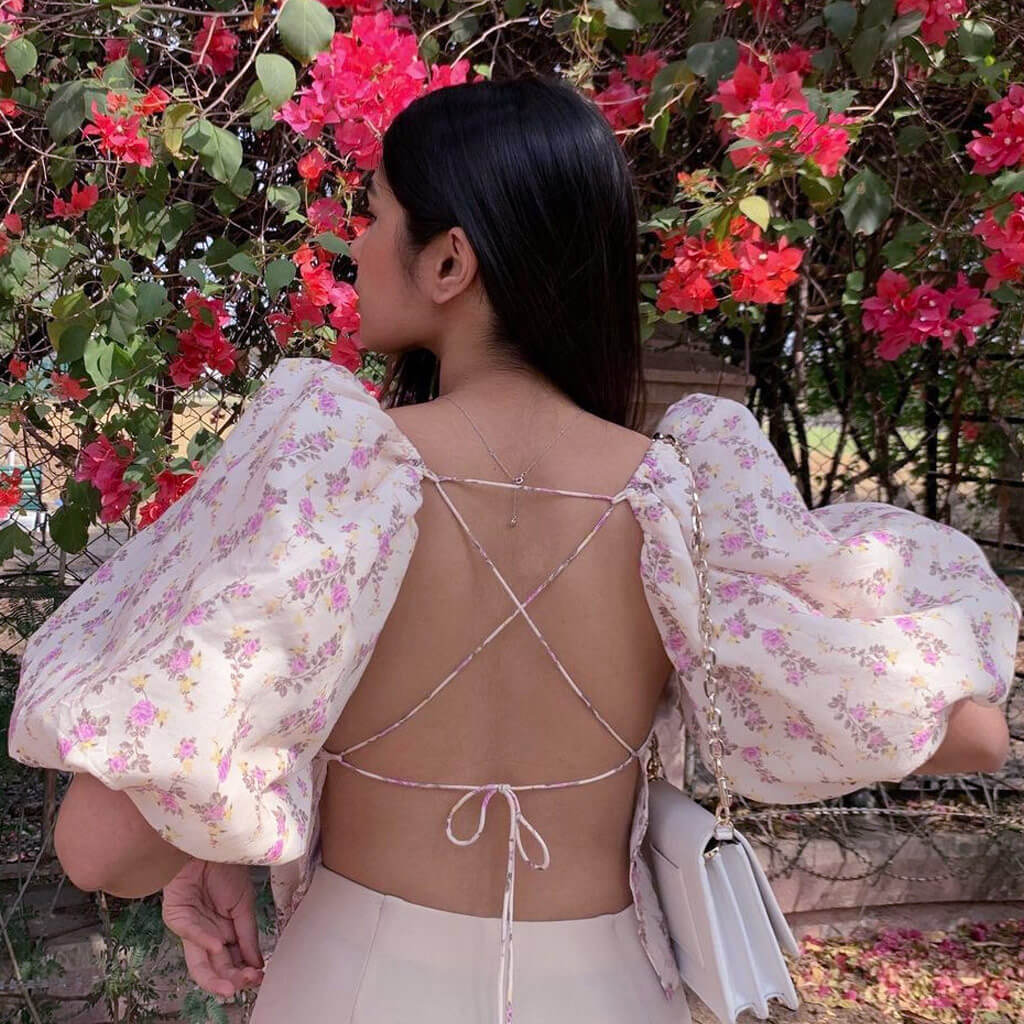 Woman in a floral print top with puff sleeves and an open back, posing in front of pink flowers.
