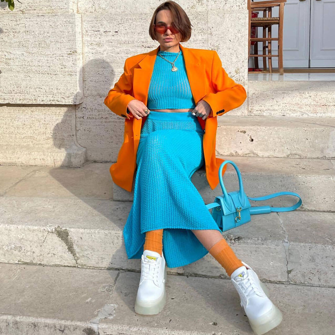 Woman sitting on stone steps wearing a blue knit crop top and skirt with an orange blazer, orange socks, and white sneakers.