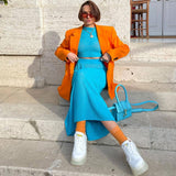 Woman sitting on stone steps wearing a blue knit crop top and skirt with an orange blazer, orange socks, and white sneakers.