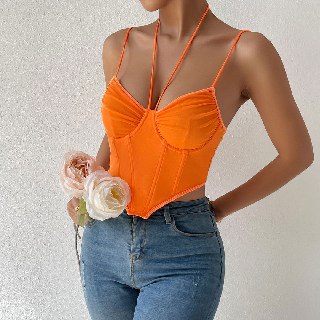 Close-up of the orange ruched mesh corset top with a plunging halter neckline and spaghetti straps, paired with ripped blue jeans. The model is holding light pink and white roses in her left hand and wearing gold rings.