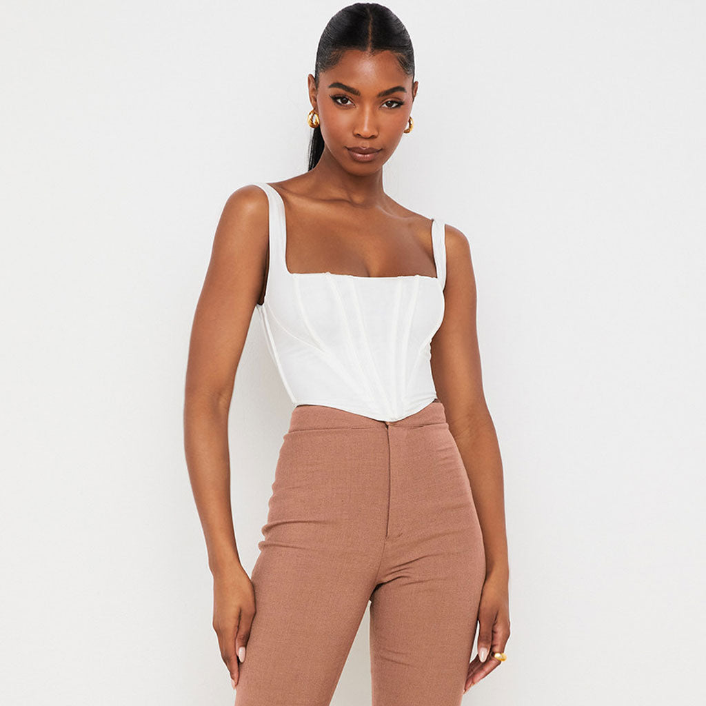 A woman poses in a white corset top and brown pants, standing against a white background.