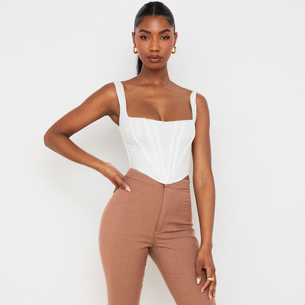A woman with her hand on her hip wears a white corset top and brown pants, against a white backdrop.