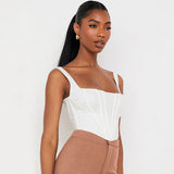 A close-up of a woman wearing a white corset top and brown pants, looking to the side against a white background.