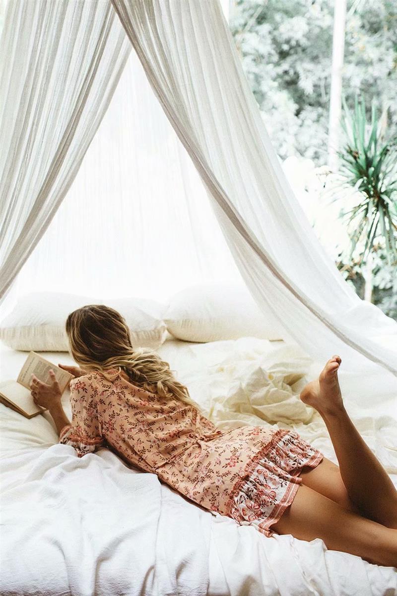 Woman in a floral peach dress lying on a bed reading a book