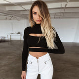 A woman wearing a black long-sleeved cropped top with a high neck and cut-out detail, paired with white pants, standing in a minimalist room.