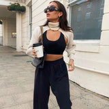 A woman wearing a cream knit cropped sweater with a high turtleneck and long sleeves layered over a black crop top and black sweatpants, holding a coffee cup and a handbag.