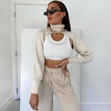 A woman wearing a cream knit cropped sweater with a high turtleneck and long sleeves layered over a white tank top and beige sweatpants, accessorized with sunglasses and gold jewelry.