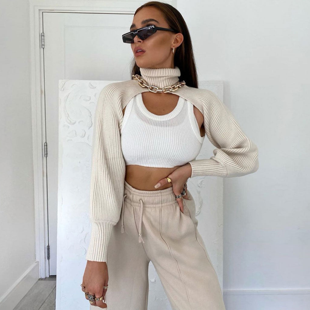 A woman wearing a cream knit cropped sweater with a high turtleneck and long sleeves layered over a white tank top and beige sweatpants, accessorized with sunglasses and gold jewelry.