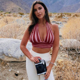 A woman with long brown hair stands in a desert landscape, holding a camera and wearing a red and white striped halter crop top and white pants.