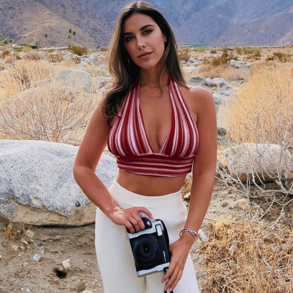 A woman with long brown hair stands in a desert landscape, holding a camera and wearing a red and white striped halter crop top and white pants.