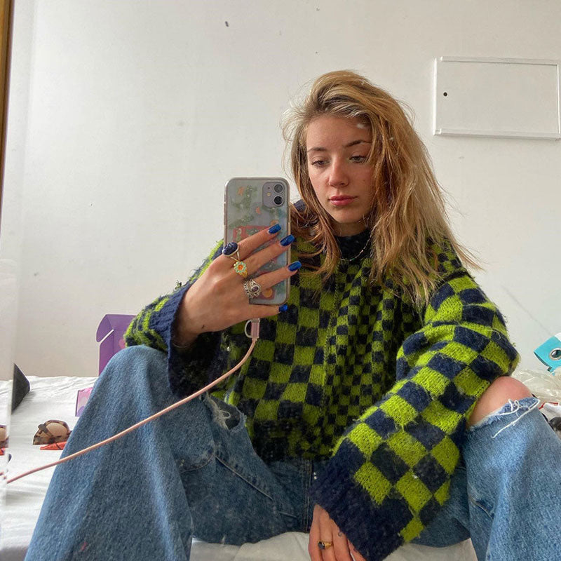 A young woman with blonde hair is sitting on a bed, holding a phone and taking a selfie. She is wearing a green and black checkerboard sweater and ripped blue jeans.