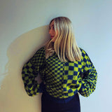A woman with blonde hair is leaning against a white wall, looking over her shoulder. She is wearing a green and black checkerboard sweater.