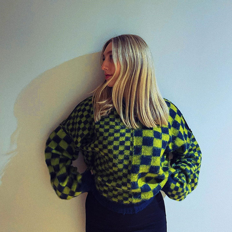 A woman with blonde hair is leaning against a white wall, looking over her shoulder. She is wearing a green and black checkerboard sweater.
