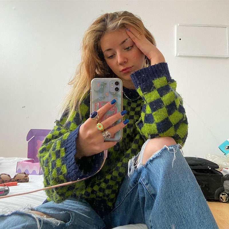 A young woman with blonde hair is sitting on a bed, holding a phone and taking a selfie. She is wearing a green and black checkerboard sweater and ripped blue jeans. She has her hand on her forehead.