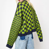 A woman with blonde hair is shown from the back, wearing a green and black checkerboard sweater and blue jeans. She is standing against a white background.