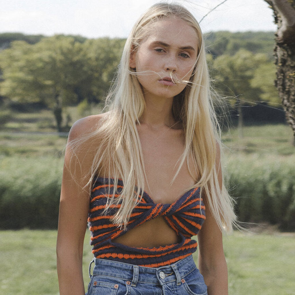 A woman with long blonde hair wearing a blue and orange striped halter top and denim jeans, standing in a grassy field.