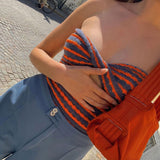 A close-up of a woman wearing a blue and orange striped halter top with a cutout, paired with blue pants and an orange bag.