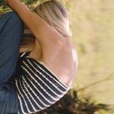 Woman from behind wearing a blue and white striped strapless top with a cut-out detail and grey pants, sitting.